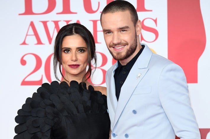 A woman in a textured black dress with a man in a light suit smile on the red carpet at a media event