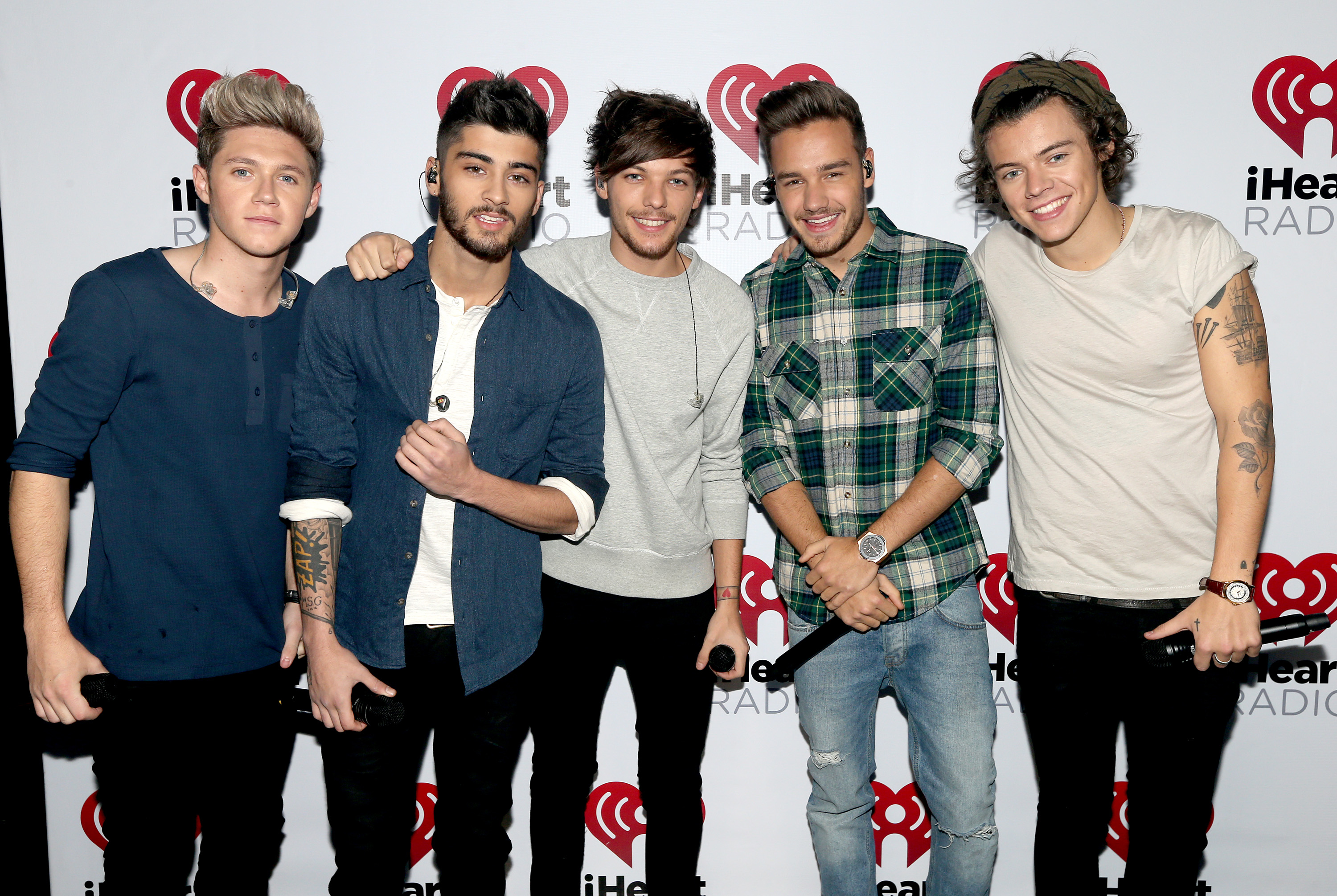 Five men on a red carpet at an iHeartRadio event, wearing casual clothing including shirts and jeans