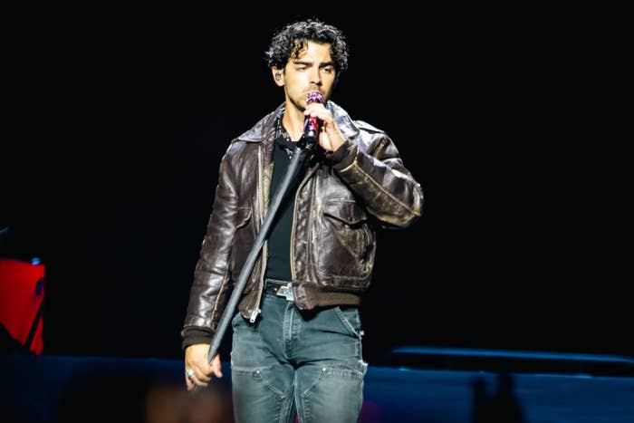 Joe Jonas performs on stage, wearing a leather jacket and jeans, singing into a microphone