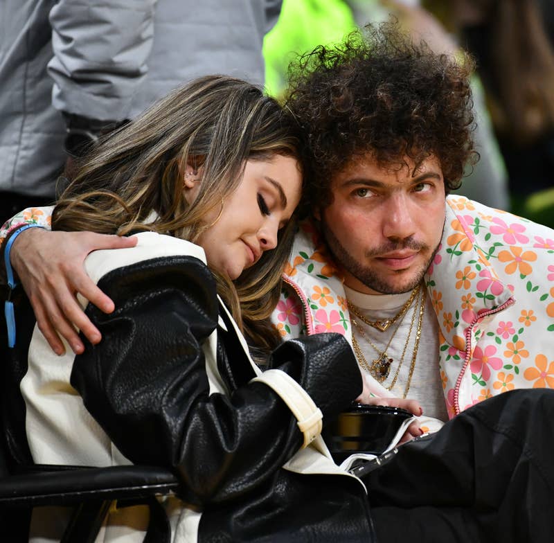 Selena Gomez and Benny Blanco attend a public event. Selena, in a black and white outfit, rests her head on Benny, who wears a floral-patterned jacket