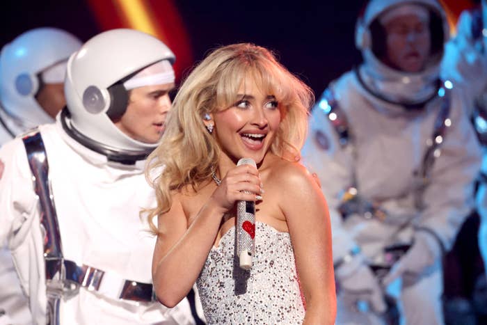 Sabrina Carpenter performs on stage wearing a sparkling dress, surrounded by dancers in astronaut costumes