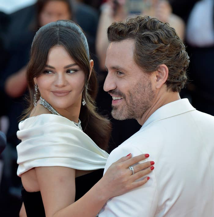 Selena Gomez and Edgar Ramírez on a red carpet event. Selena is wearing a chic off-shoulder gown with sparkling earrings. Edgar is in a suit
