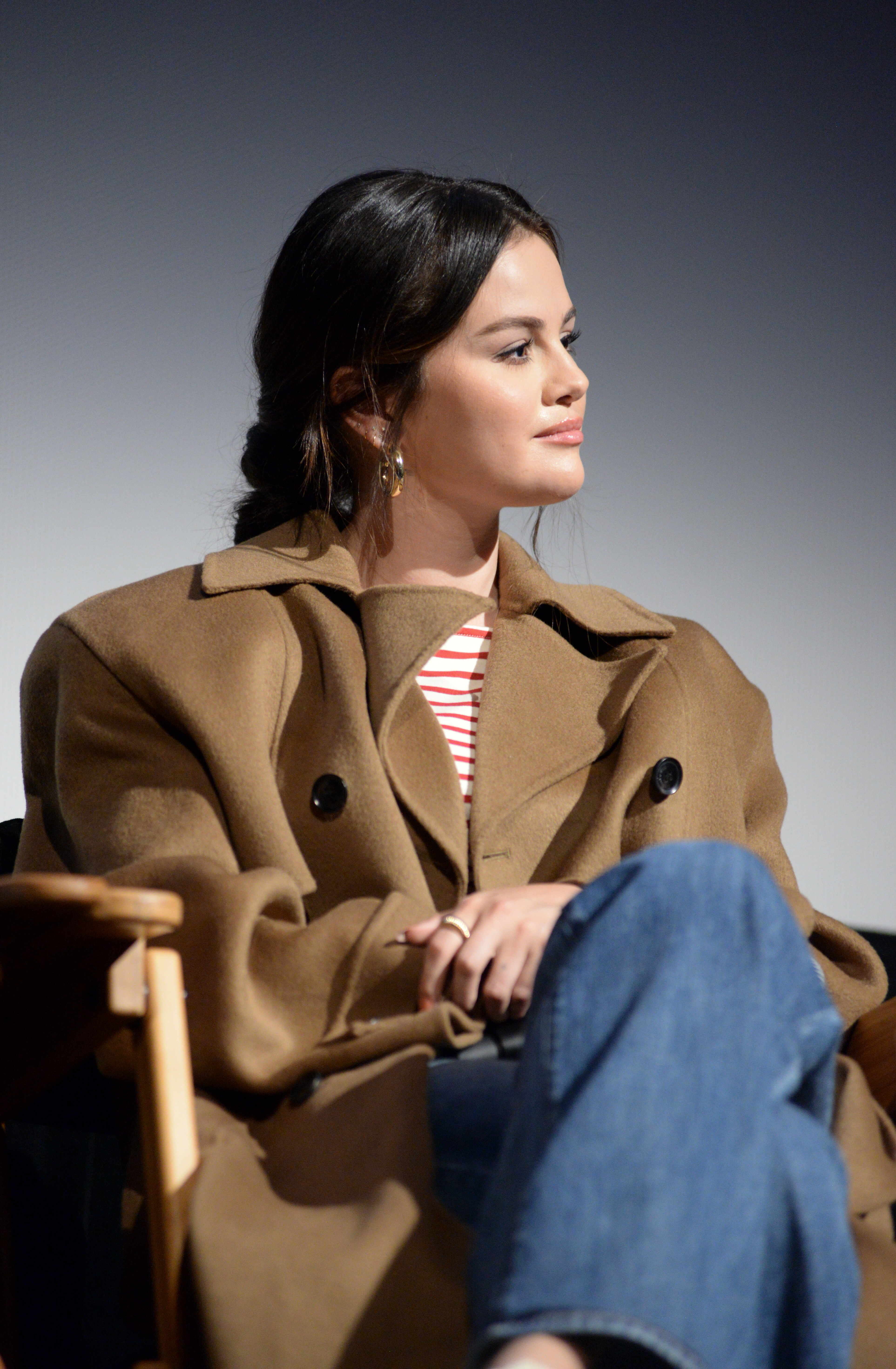 Selena Gomez sits and listens attentively, wearing a brown coat over a striped shirt and blue jeans