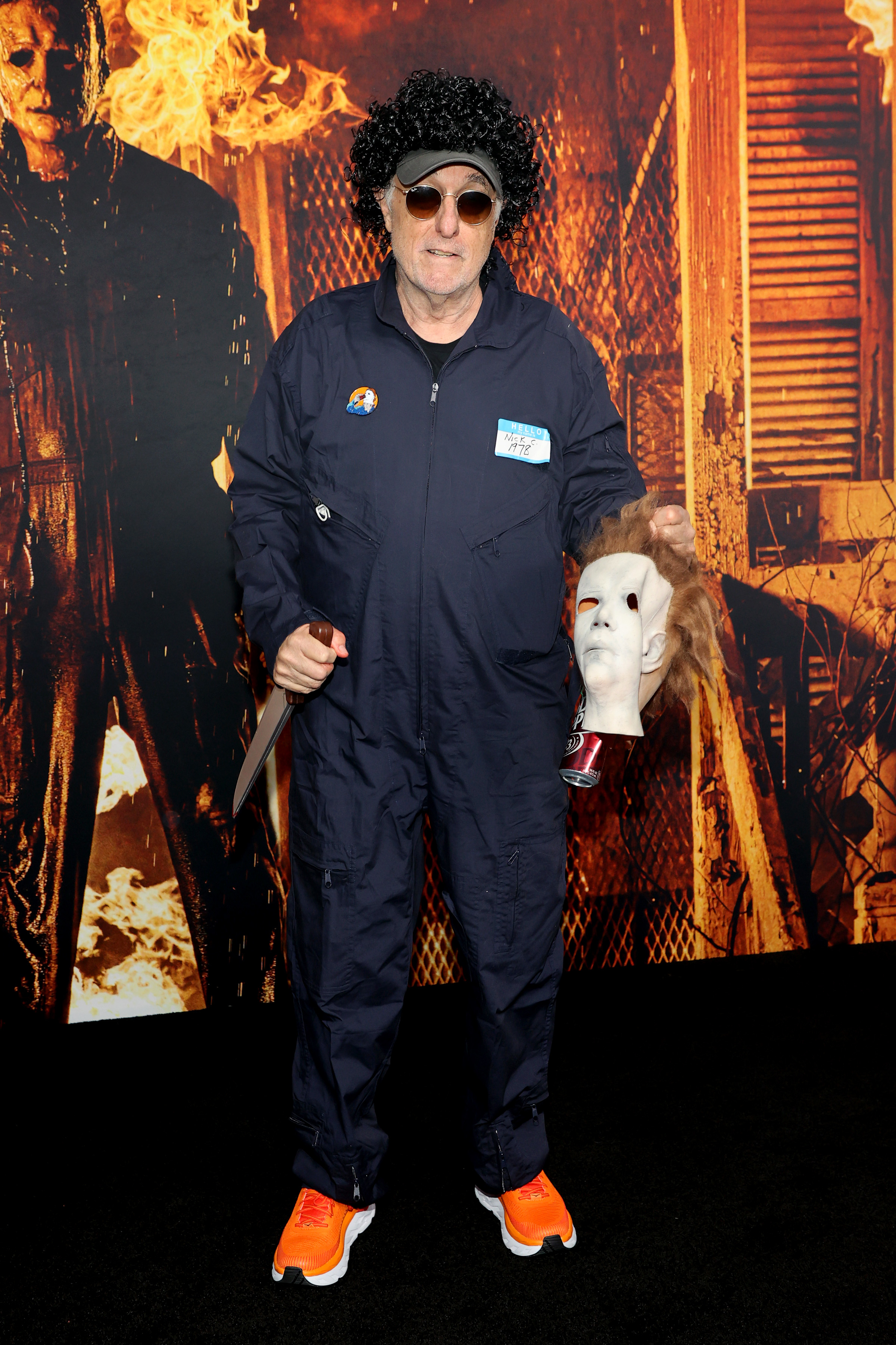 Nick Castle wearing a blue jumpsuit and orange sneakers while holding a faux knife and Michael Myers mask at the premiere of "Halloween Kills"