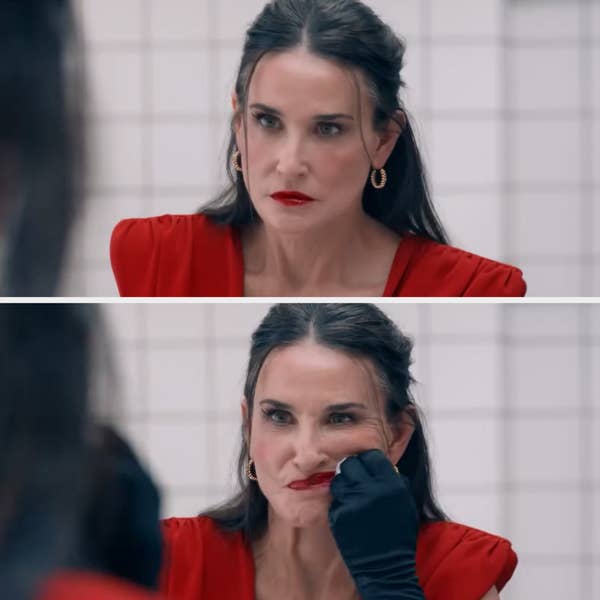 Demi Moore, wearing a chic red dress and gold hoop earrings, looks worried in a bathroom mirror in this scene from "Brave New World."