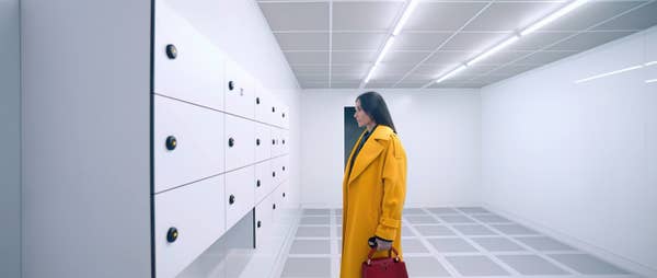 Woman in a stylish trench coat looks at minimalist wall storage units in a modern, brightly lit room. She holds a handbag. Names are unknown