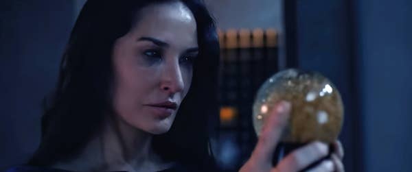 A person with long dark hair stares intensely at a glowing orb in their hand, set against a dimly lit background