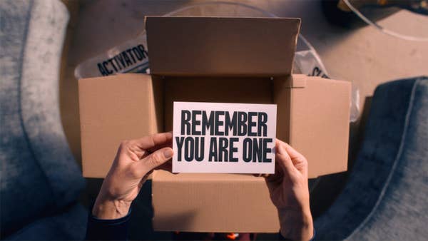 A person holds a card with the text &quot;REMEMBER YOU ARE ONE&quot; over an open cardboard box