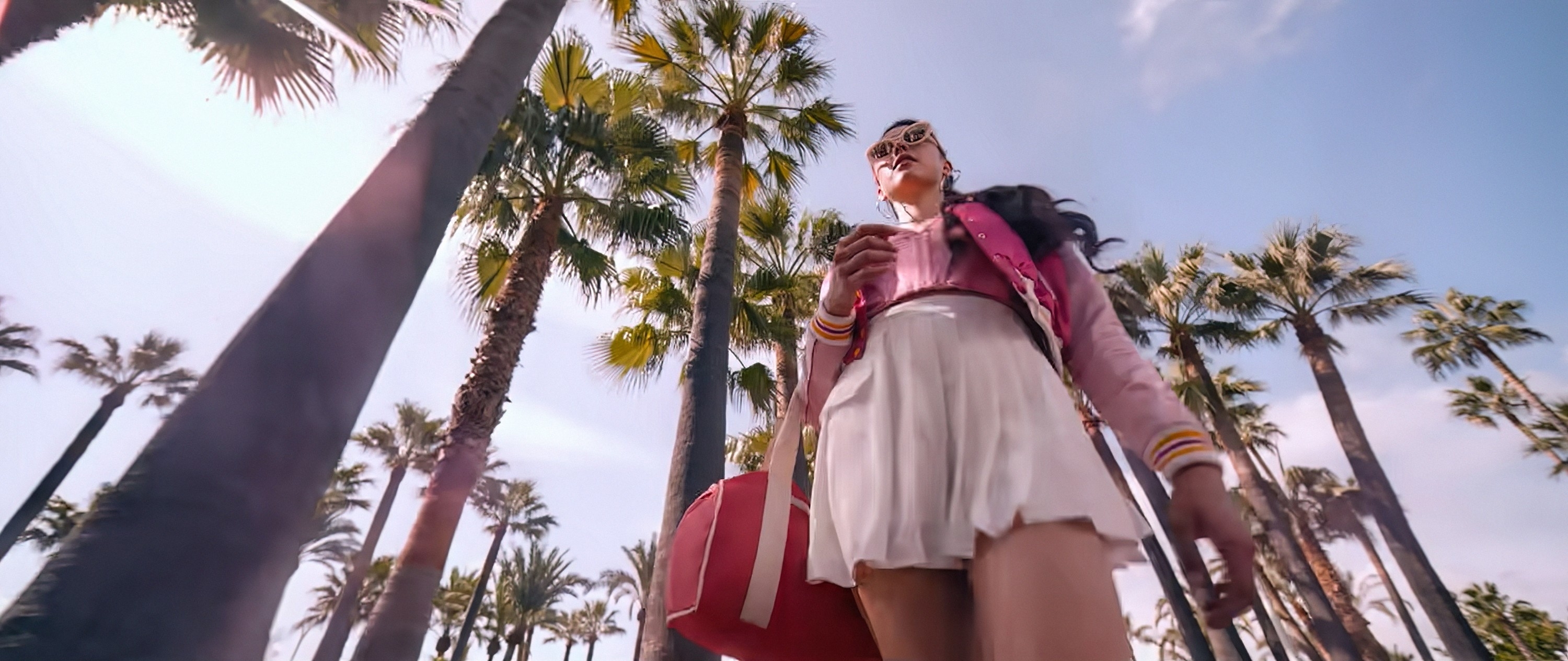 A person wearing sunglasses, a pink jacket, and a white skirt is walking among tall palm trees on a sunny day, holding a drink and bag. Names unknown