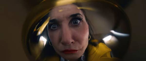 Close-up of a woman with wide eyes and a surprised expression, looking through a distorted lens. The woman is wearing a yellow garment