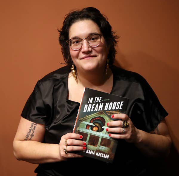 Carmen Maria holding her book &quot;In the Dream House&quot;, wearing glasses, a black satin blouse, with short hair