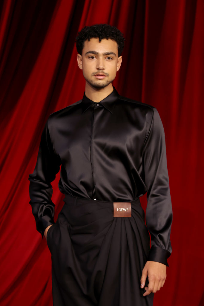 A person stands on a red carpet wearing a sleek, dark satin shirt and flowing high-waisted pants, with a Loewe belt detail