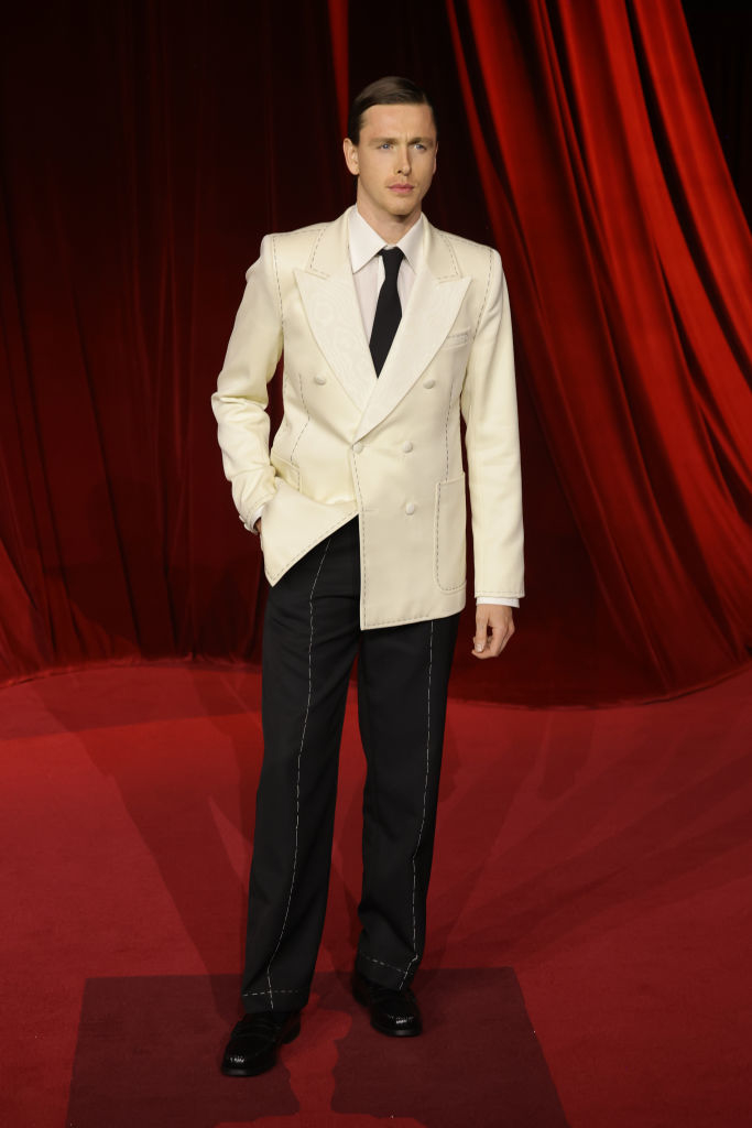 A man in a white double-breasted suit and black pants stands on a red carpet