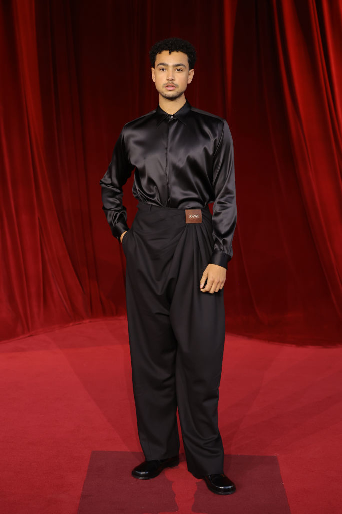 A person stands on a red carpet wearing a black satin shirt and high-waisted trousers