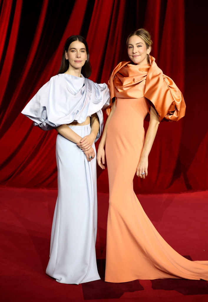 Two women pose on a red carpet in elegant gowns with large, draped sleeves