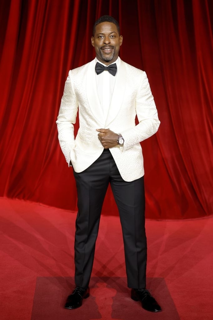 Man in a white textured tuxedo jacket, black bow tie, and black pants on a red carpet, smiling with hands in pockets