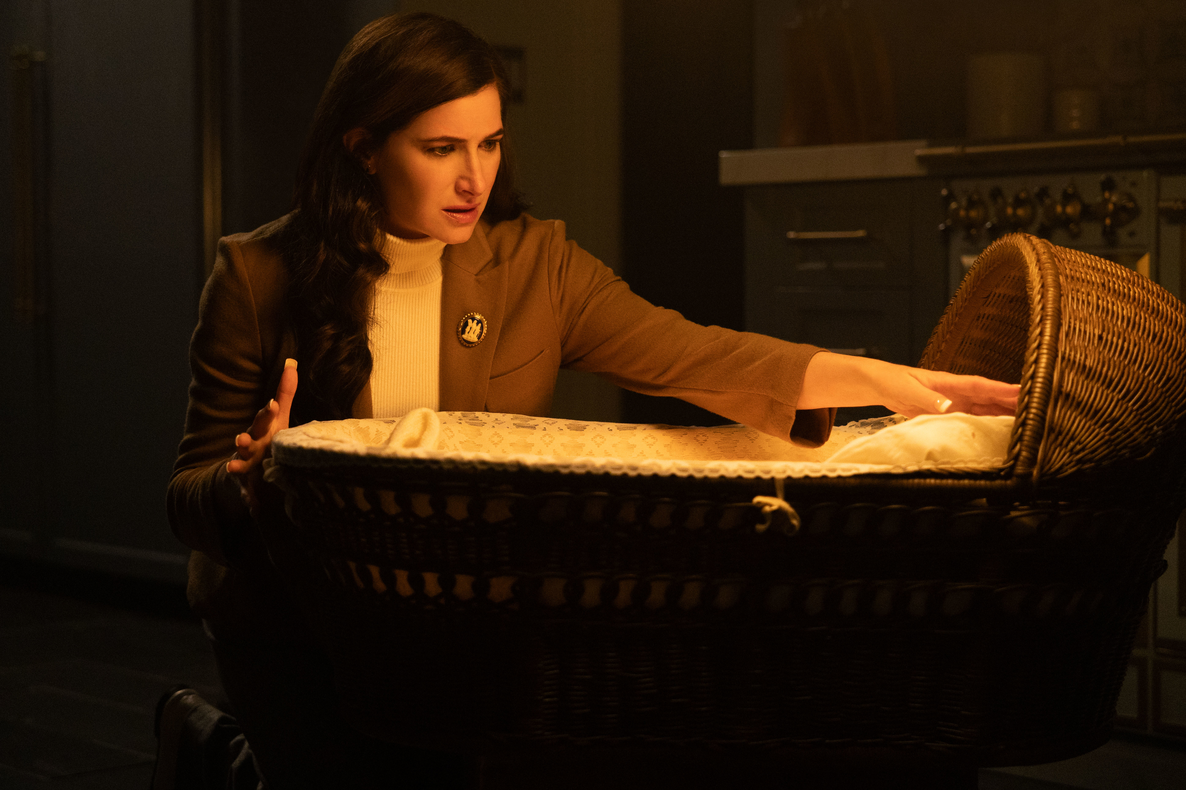 A woman with long dark hair in a brown blazer reaches into a wicker bassinet with a concerned expression