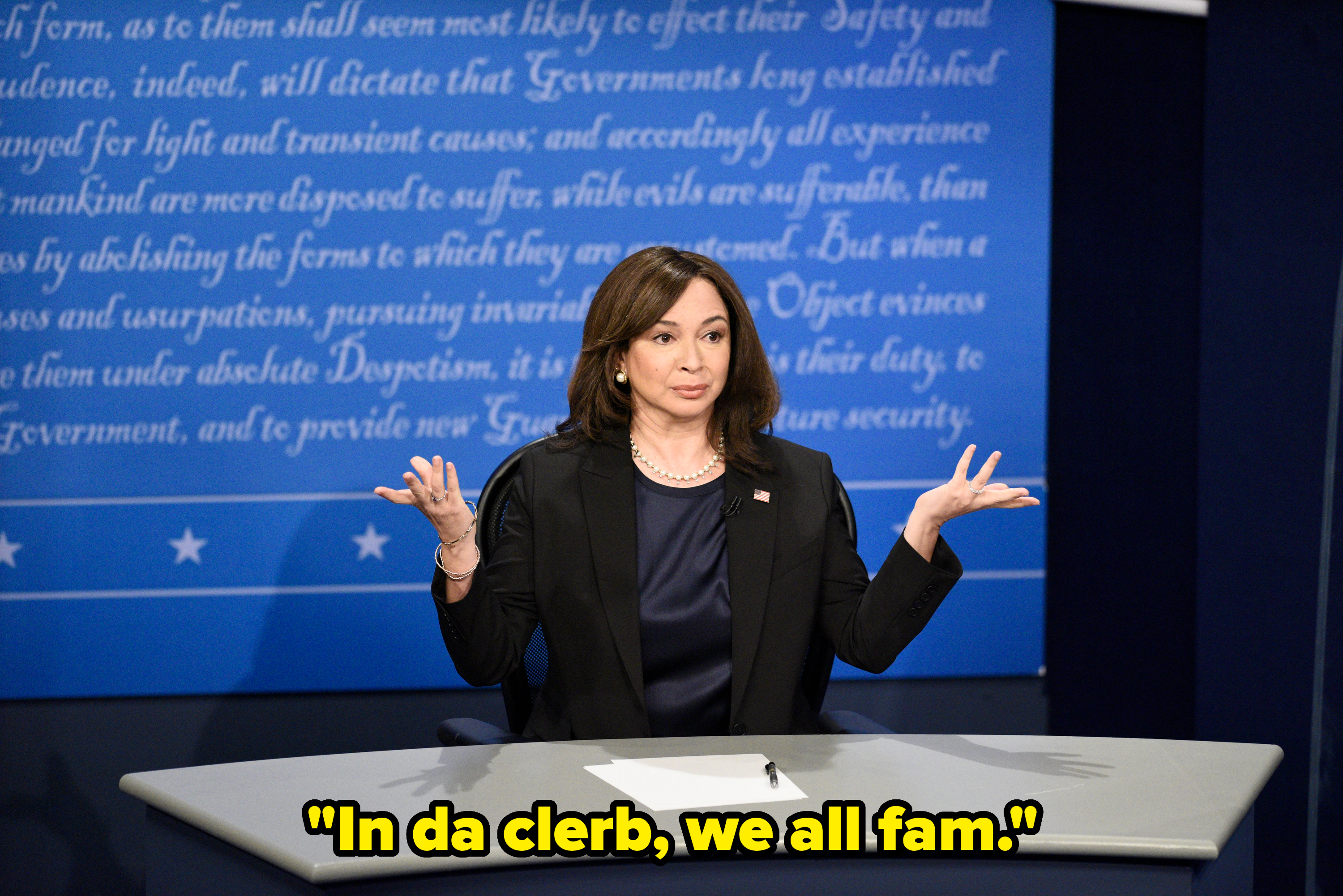 Maya Rudolph as Kamala Harris sits at a debate desk, gesturing with both hands