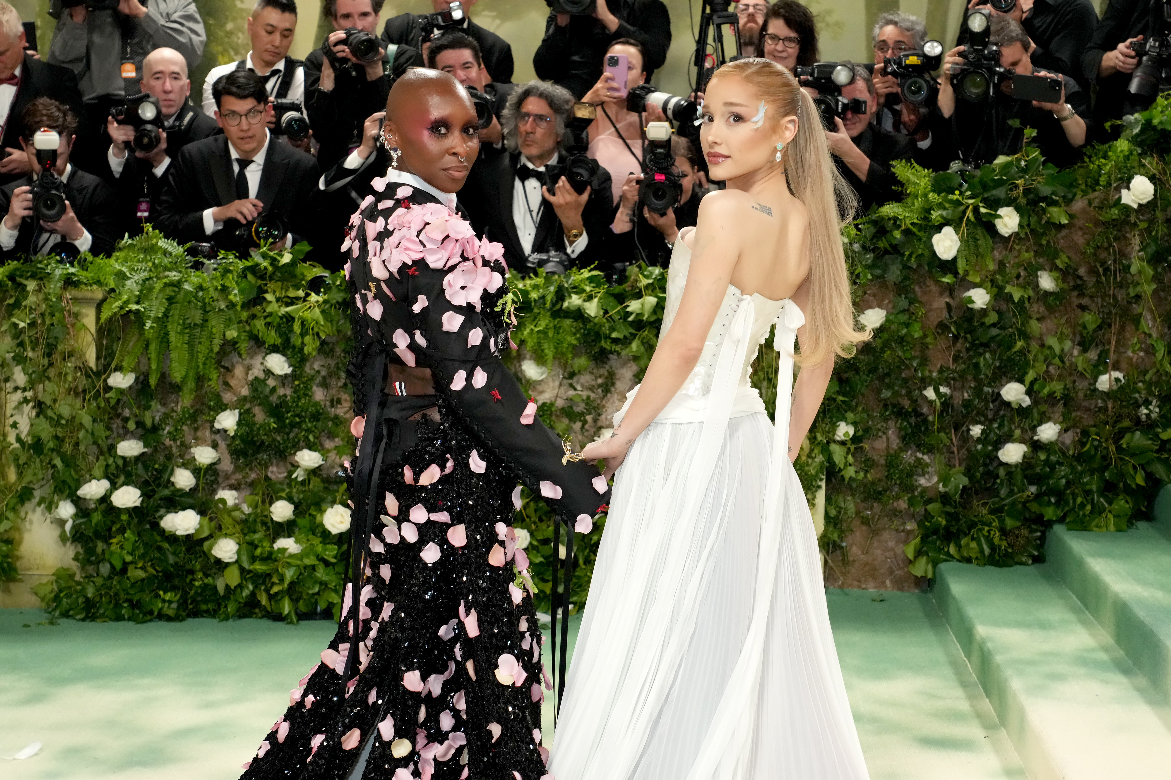 Two people on a red carpet, one in a floral gown and the other in a strapless white dress, facing photographers and holding hands