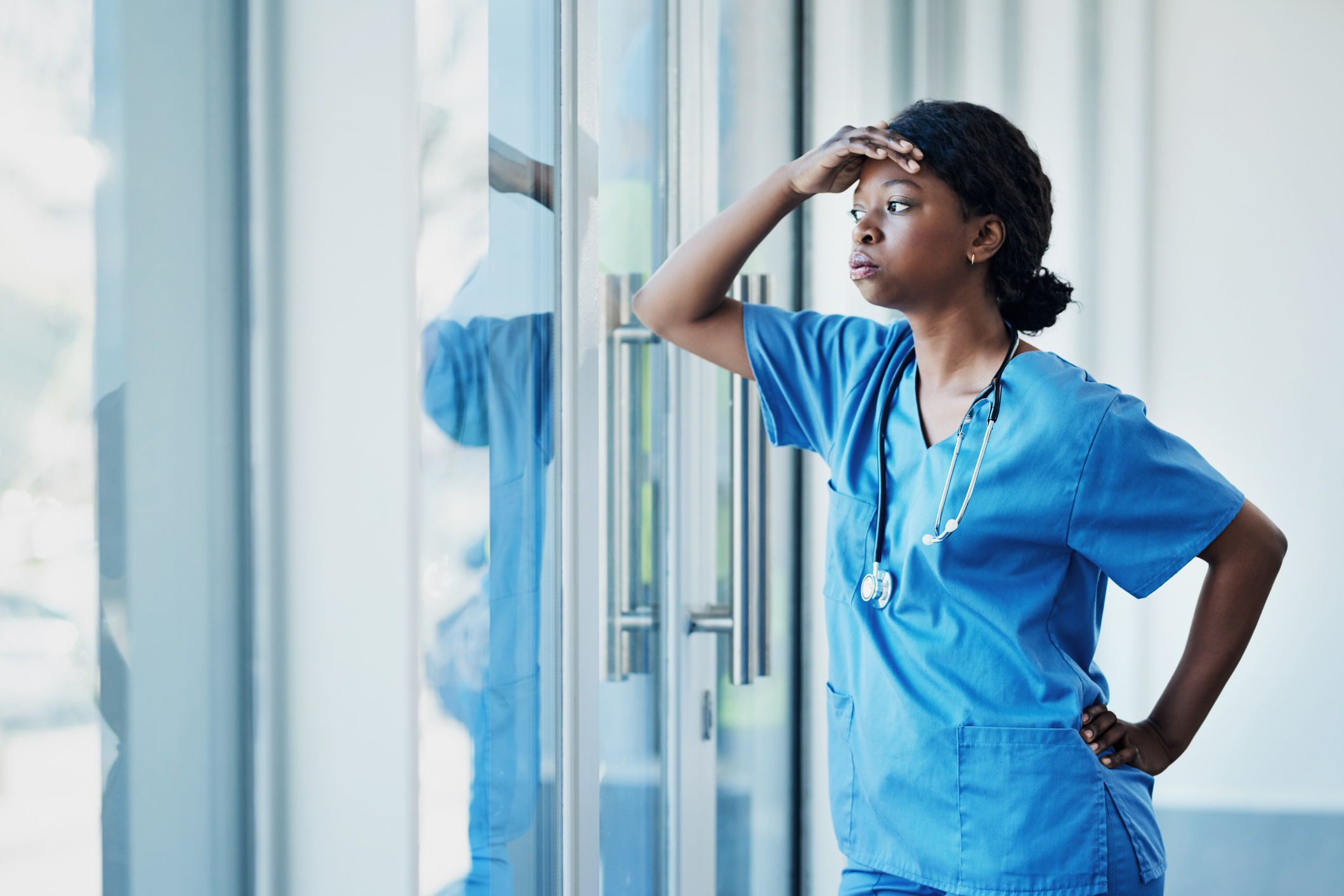 A healthcare worker in scrubs looks out a window with a stethoscope around their neck, appearing thoughtful