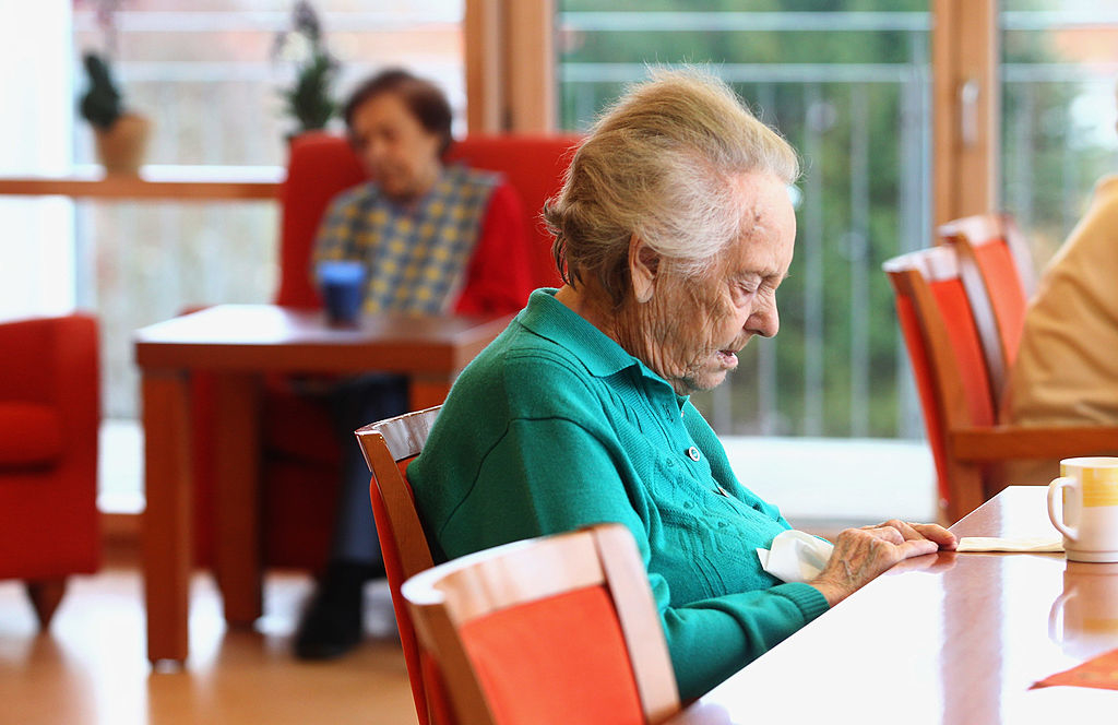 Elderly woman sitting at a table in a senior living facility, appearing contemplative, with another person blurred in the background