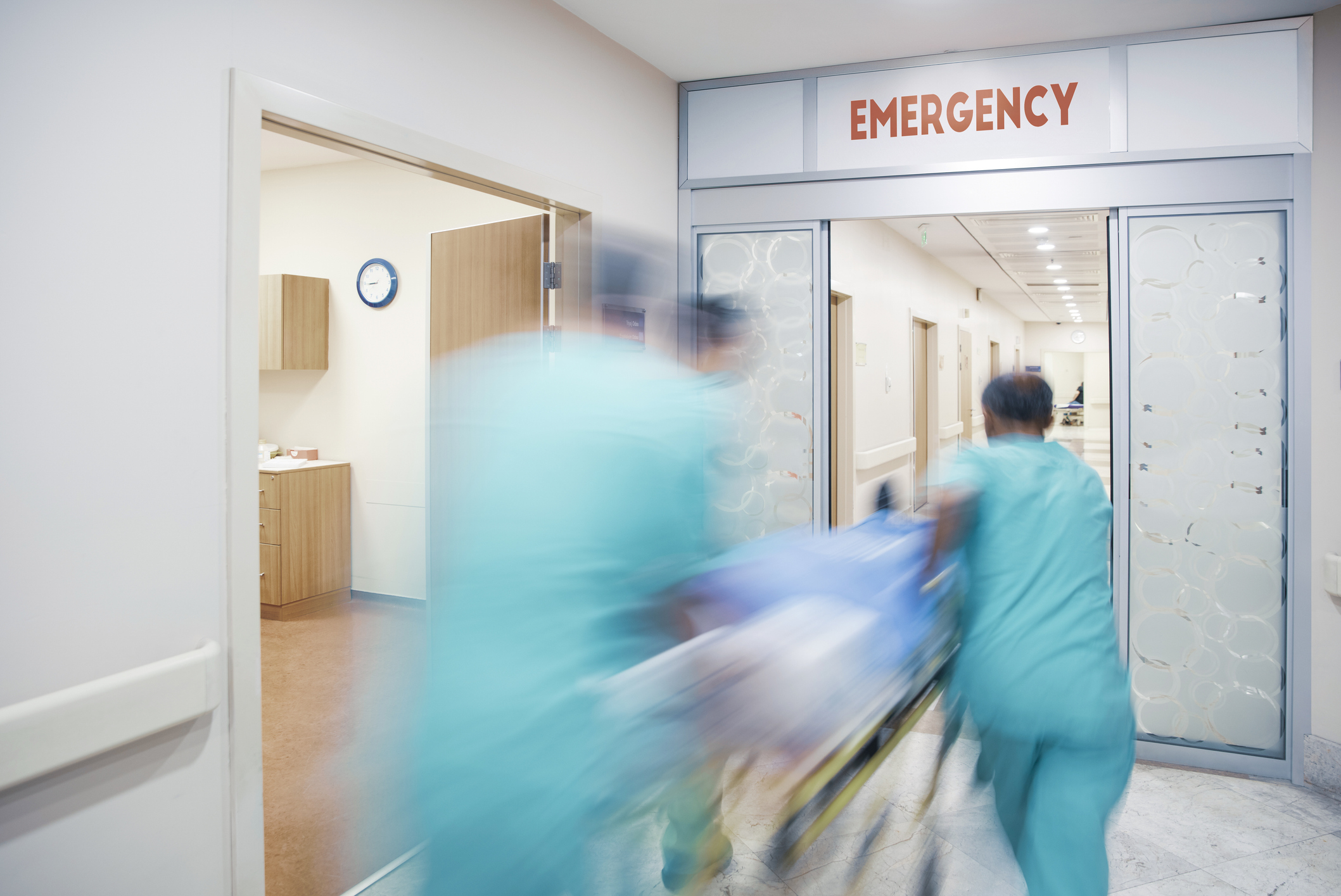 Blurred medical staff rush a patient on a gurney through hospital doors marked &quot;Emergency.&quot;