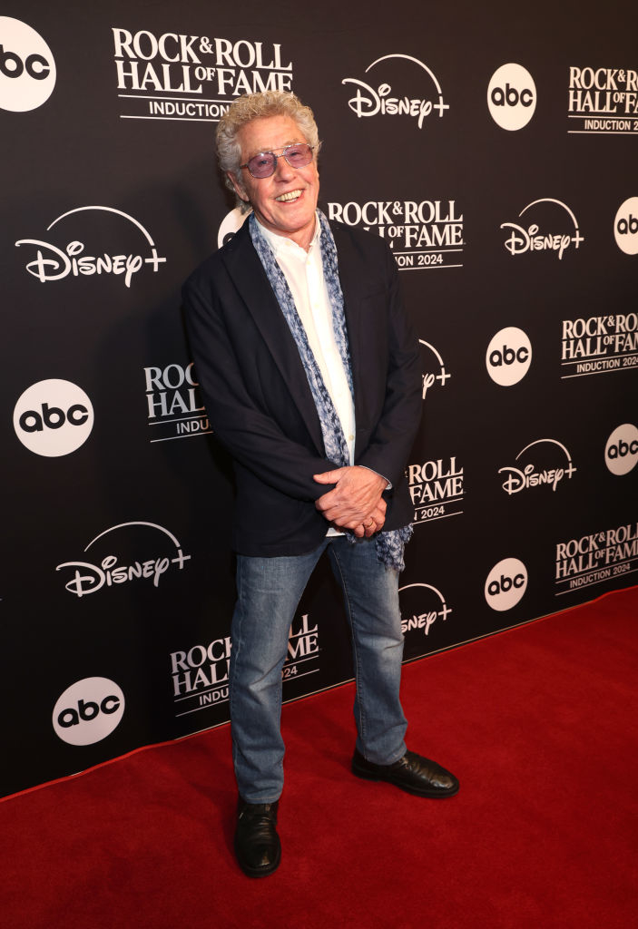 A person on a red carpet wearing jeans, a white shirt, and a blazer with a scarf at a Rock &amp;amp; Roll Hall of Fame event