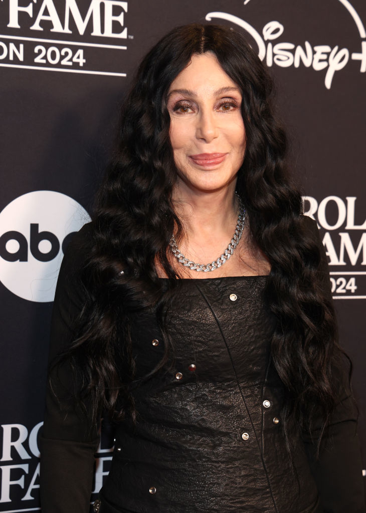 I don’t know who this is, but the image shows a person on a red carpet wearing a textured black outfit with long hair and a chain necklace