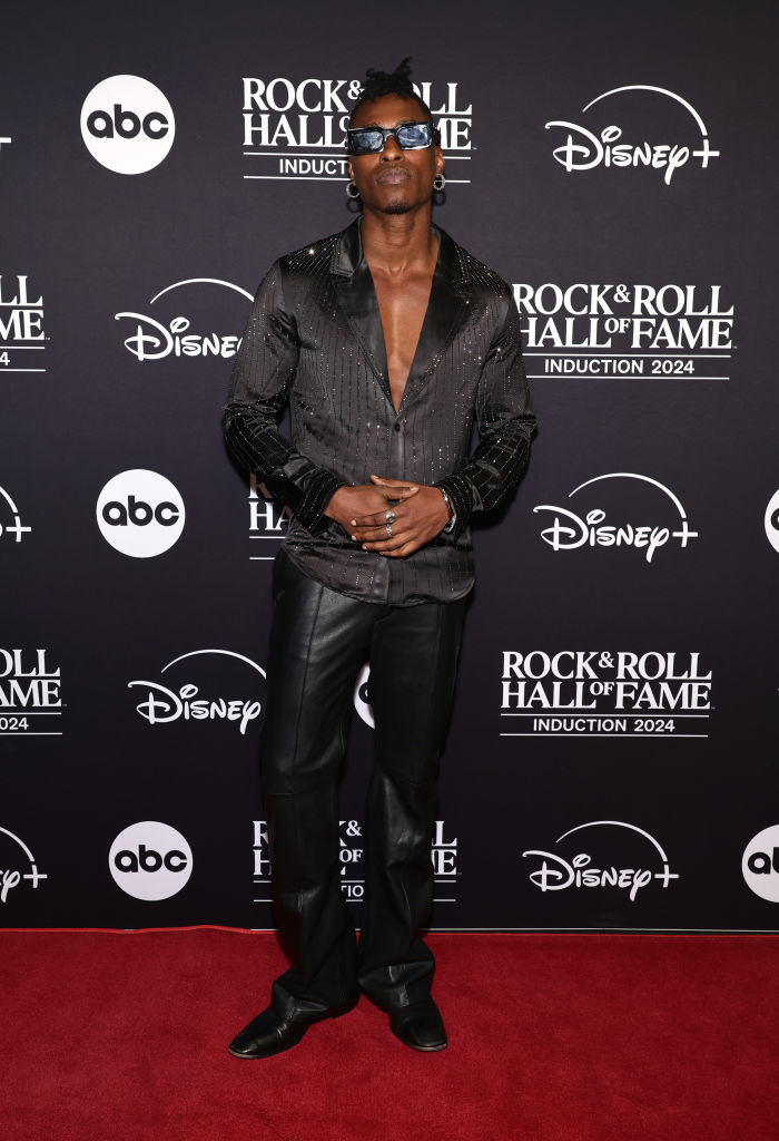 Person on a red carpet in a shiny, open black shirt and leather pants at the Rock &amp;amp; Roll Hall of Fame Induction 2024 backdrop