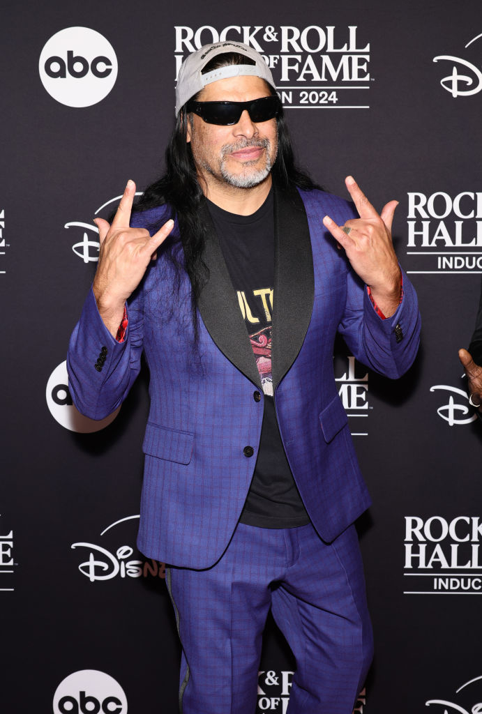 A man in a purple suit and sunglasses poses on a red carpet. Background includes logos for ABC, Disney, and the Rock &amp;amp; Roll Hall of Fame Induction 2024