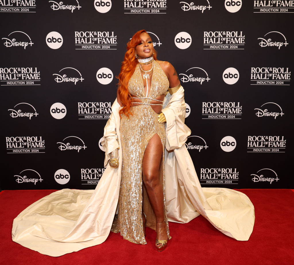 I don’t know who is in the image. The person is wearing a shimmering gown with a high slit and a long coat, standing on a red carpet with a backdrop