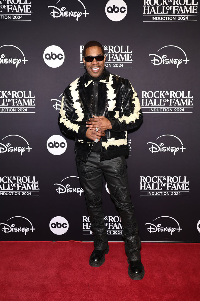 A person in stylish leather attire with a textured jacket poses on a red carpet at a Rock &amp;amp; Roll Hall of Fame event backdrop