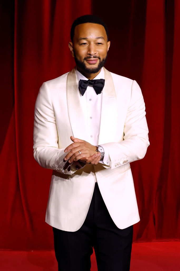 A person in a white suit with a black bow tie stands in front of a red curtain on a red carpet