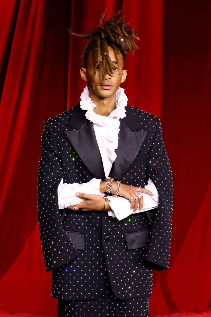 A person on a red carpet wearing a studded blazer and a ruffled shirt