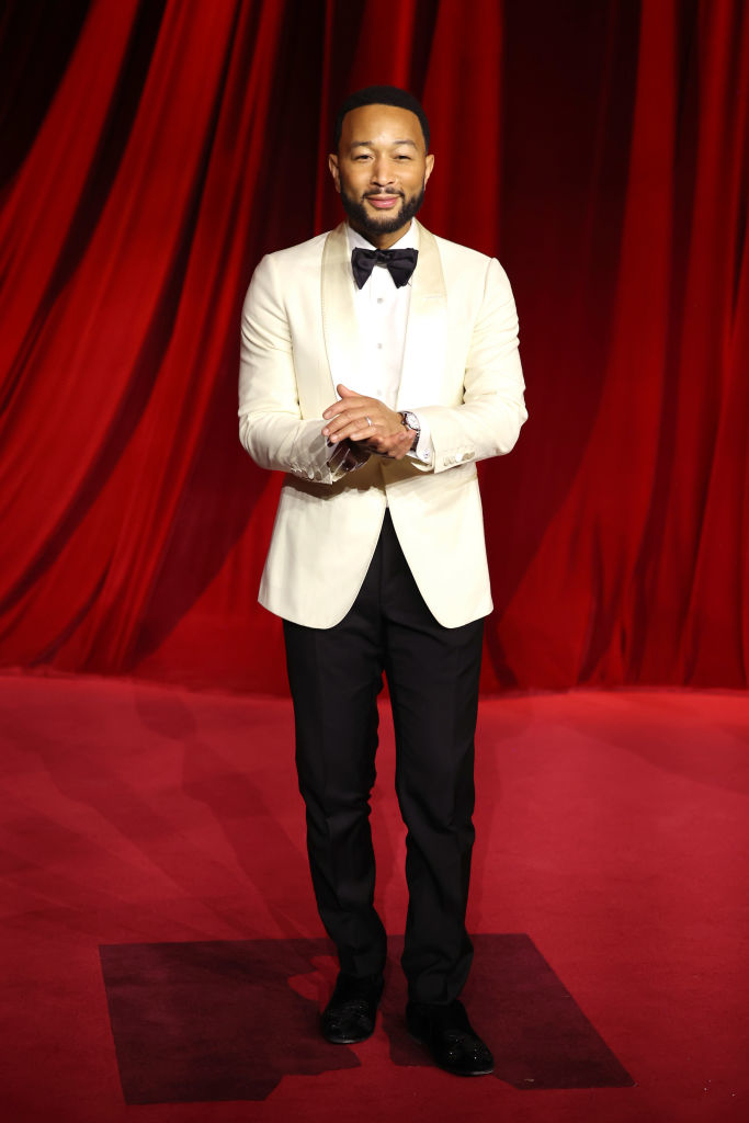 A person in a white tuxedo jacket and black pants stands on a red carpet backdrop