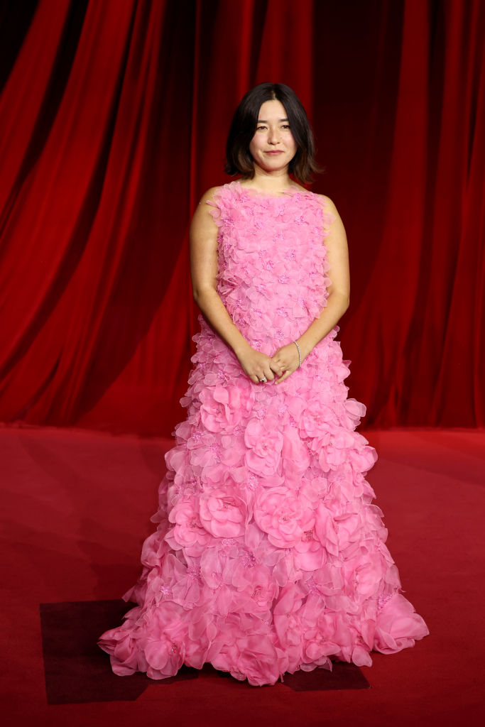 A woman in a textured, floral pink gown stands on a red carpet