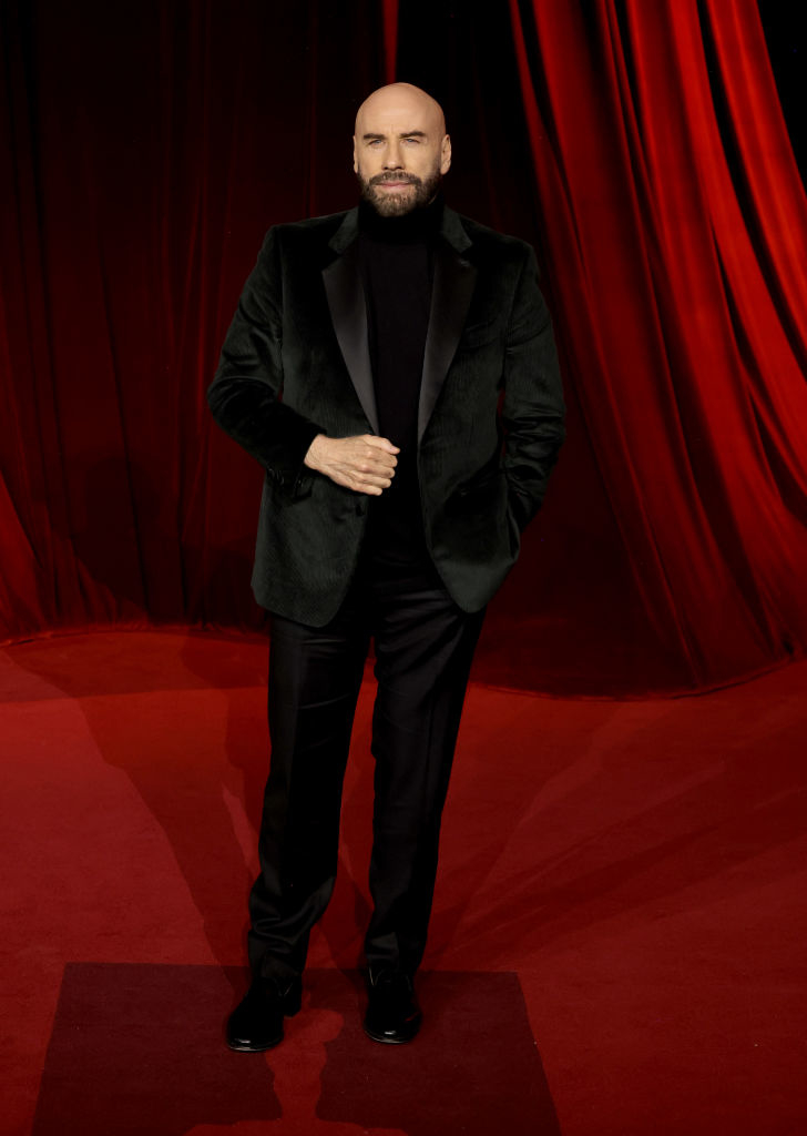 A person in a black suit stands on a red carpet, posing confidently