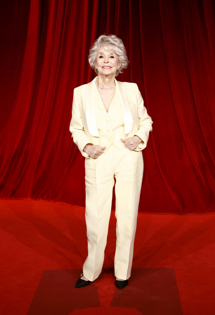 An older woman in a tailored, light-colored suit stands on a red carpet with a rich red curtain backdrop, smiling confidently