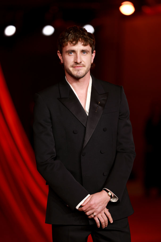 A man in a tailored black suit with a white shirt stands on a red carpet