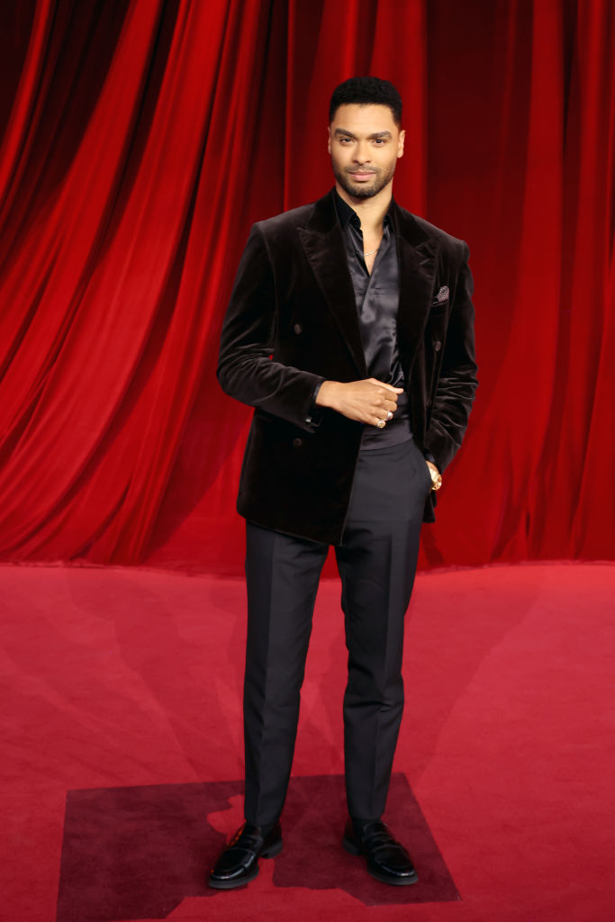 A person stands on a red carpet in a velvet suit, dress shirt, and dress shoes with a backdrop of flowing red fabric