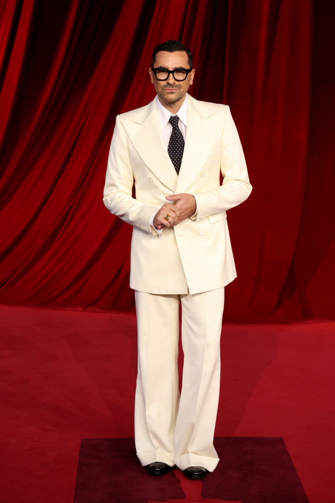 A person in a white suit with wide-legged pants and black glasses stands on a red carpet in front of a red curtain backdrop