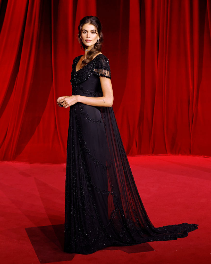 A person in an elegant, flowing gown with beaded details poses on a red carpet with a red curtain backdrop