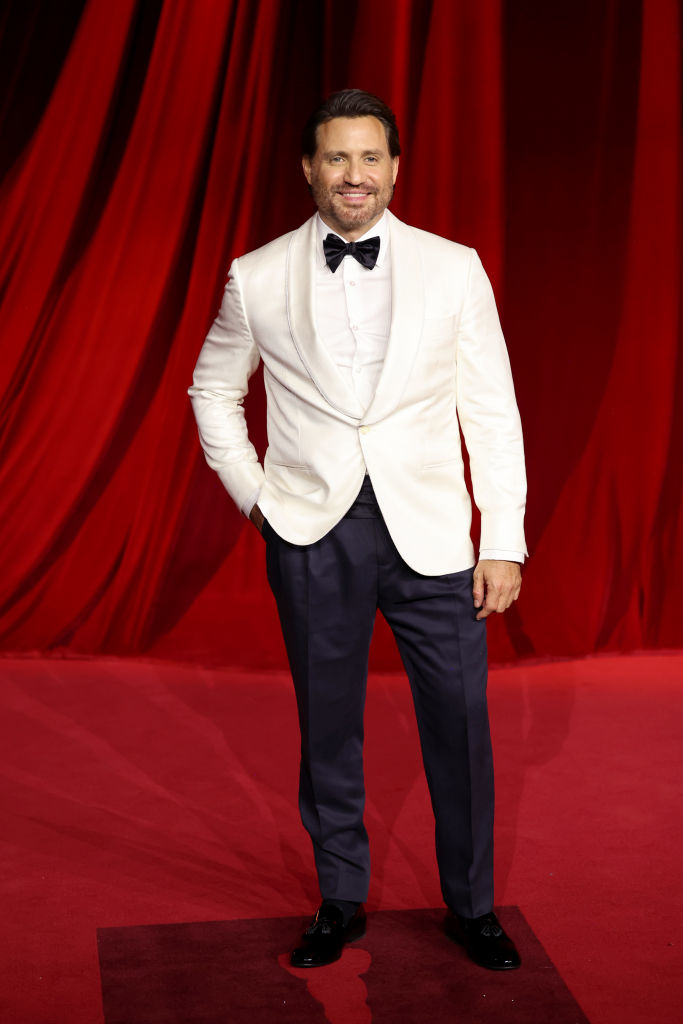 A man in a white blazer and black pants stands on a red carpet