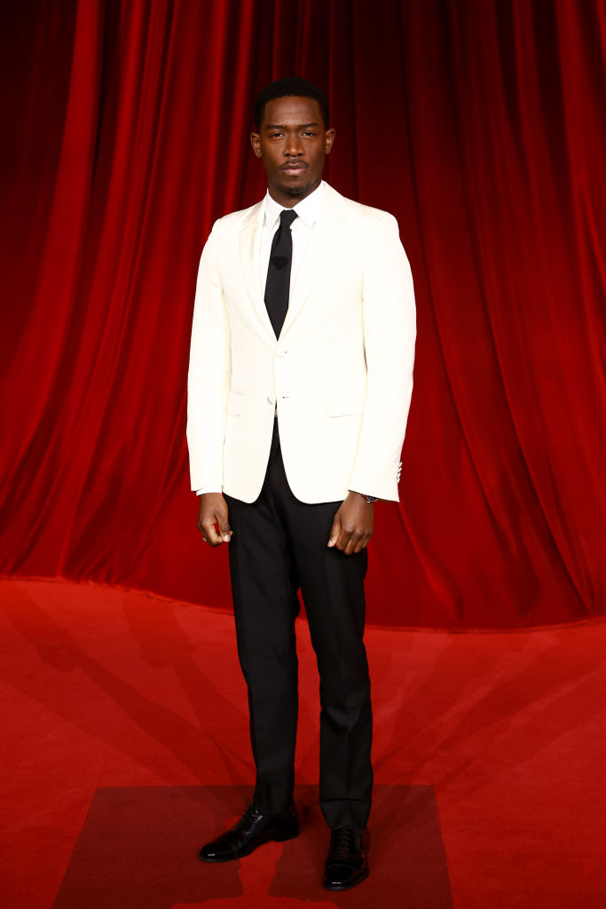 A person stands on a red carpet wearing a white blazer, black pants, and black shoes