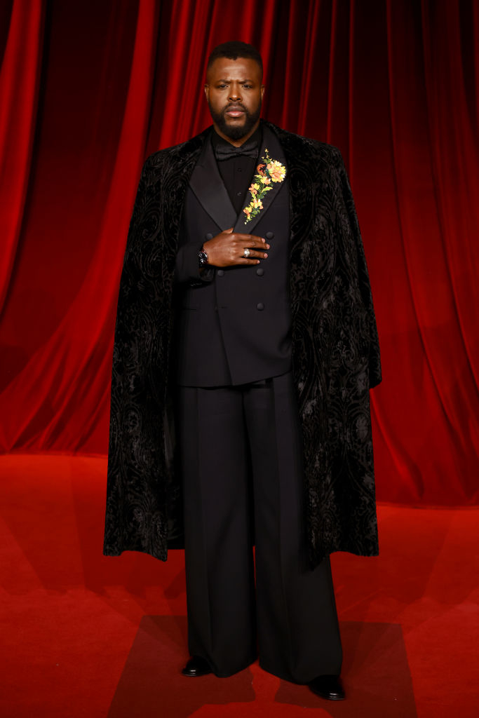 A person stands on a red carpet wearing an elegant black suit with a velvet cape and floral brooch