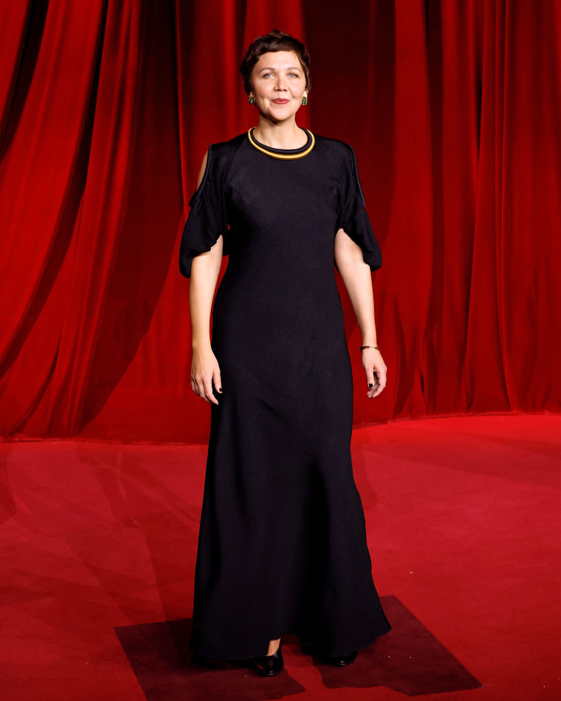 A woman in a black gown with gold jewelry stands on a red carpet with a backdrop of red curtains