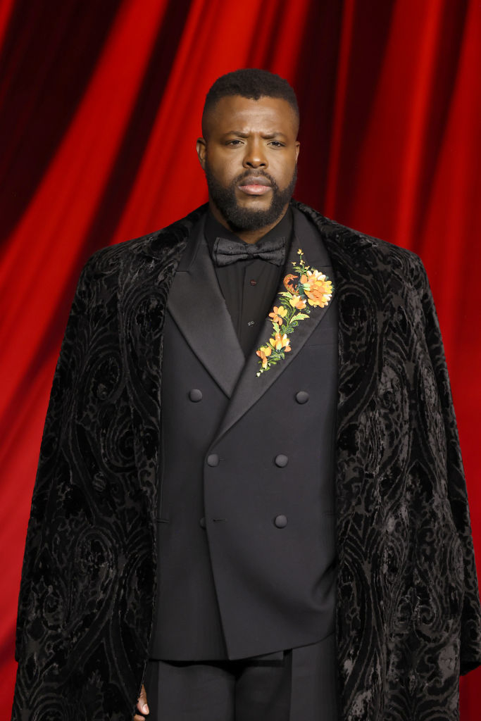 A person in a black suit with floral embroidery and a velvet cape stands against a red background