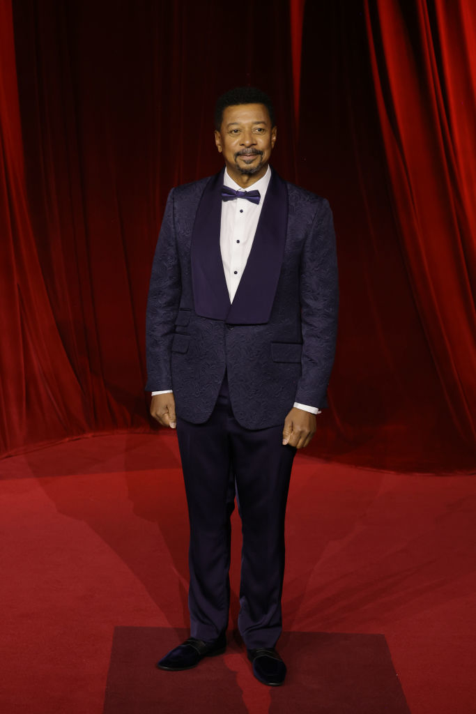 A person in a tuxedo with a bow tie stands on a red carpet with a velvet backdrop