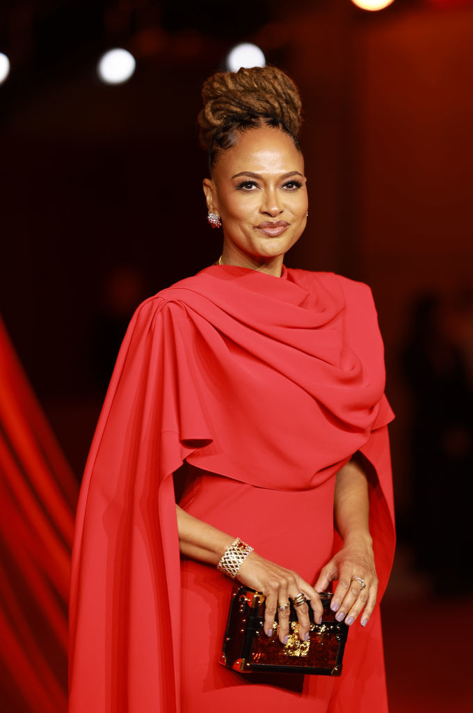 A woman on a red carpet wearing an elegant gown with draped fabric and holding a clutch purse, accessorized with bracelets and earrings