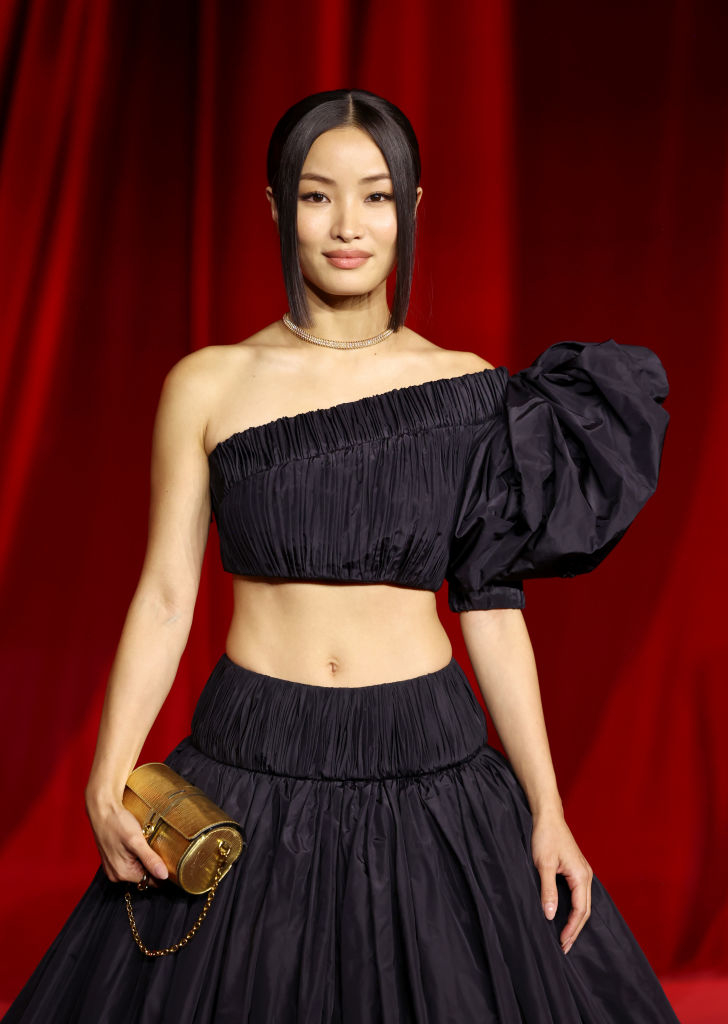 Anna Sawai in a stylish asymmetric black gown on a red carpet holds a gold clutch, with hair up and minimal jewelry